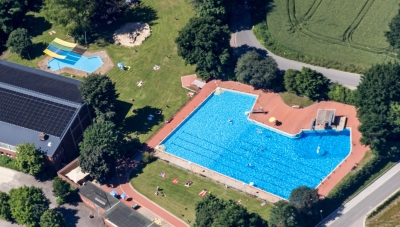 Blick von oben auf das Gelände des Freibads Mettingen. Große Rasenflächen und zwei Schwimmbereiche. Umrahmt von großen Bäumen. Links ist das Hallenbad zu sehen.