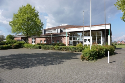 Außenansicht auf die Tüötten-Sporthalle Mettingen. Im Vordergrund der Parkplatz und eine niedrige Hecke. Im Hintergrund die Sporthalle und ein Baum.