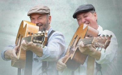 Das Duo hotte&bassmass hält ihre Gitarren in die Kamera und zielt in Blickrichtung. Sie lächeln.