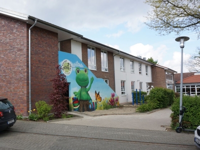 Blick von der Straße auf die Paul-Gerhard-Schule Mettingen. Rote Ziegelsteine und eine bemalte Hauswand mit einem winkenden Frosch als Motiv.