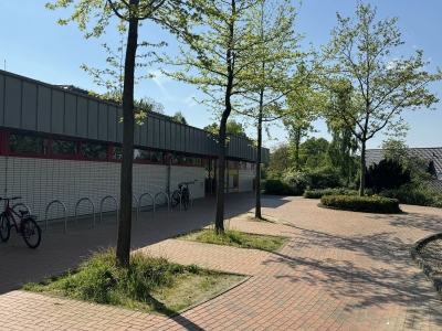 Außenansicht auf das Hallenbad Mettingen. Fahrradständer und ein gepflasterter Gehweg. Im Hintergrund das Hallenbad Mettingen