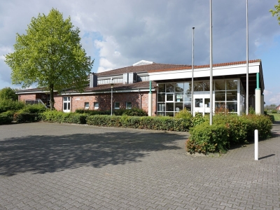 Außenansicht auf die Tüötten-Sporthalle Mettingen. Im Vordergrund der Parkplatz und eine niedrige Hecke. Im Hintergrund die Sporthalle und ein Baum.