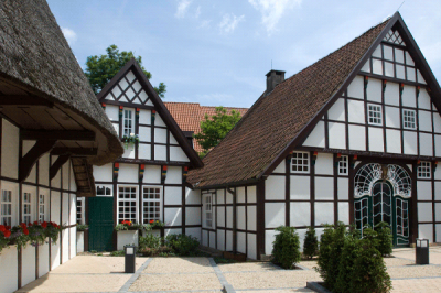 Außenansicht auf schöne Fachwerkhäuser mit weißer Front, roten Blumen und hohen Dächern. Die Sonne scheint.