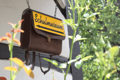 Eine alte Schultasche hängt an einer Hauswand. An ihr ist ein gelbes Schild mit der Aufschrift 