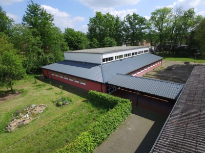 Außenansicht in der Vogelperspektive auf die Sporthalle Josef-Schule Mettingen. Rote Klinkerwände umgeben von Rasen und Bäumen.